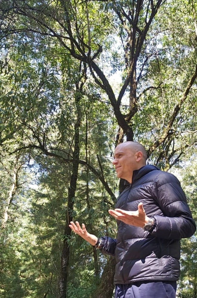 Man in black puffer jacket gesturing while speaking in a forest of tall green trees